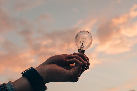 Silhouette of a hand holding a light bulb against a vibrant sunset sky, symbolizing ideas and innovation.